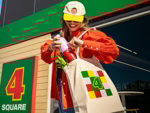 Pretty Girl walking out of Four Square with groceries in a four square branded tote bag and drink bottle.